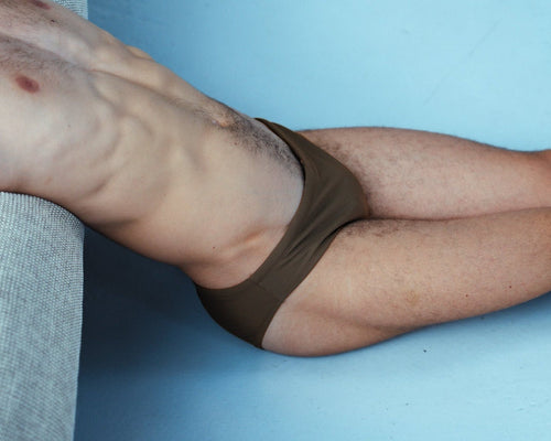A close-up lifestyle shot of a man's lower torso and legs wearing brown briefs, lying next to a gray textured cushion.