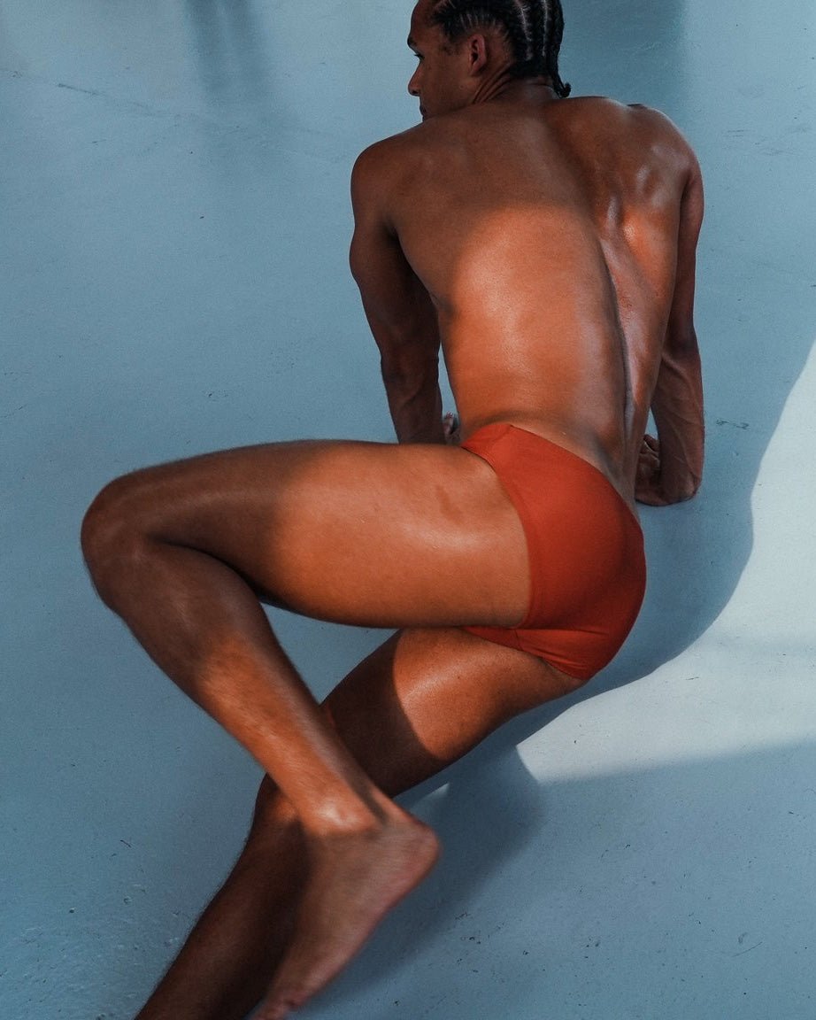 A man wearing bright orange-red briefs sits on a light blue floor, viewed from behind with strong sunlight casting shadows on the surface.