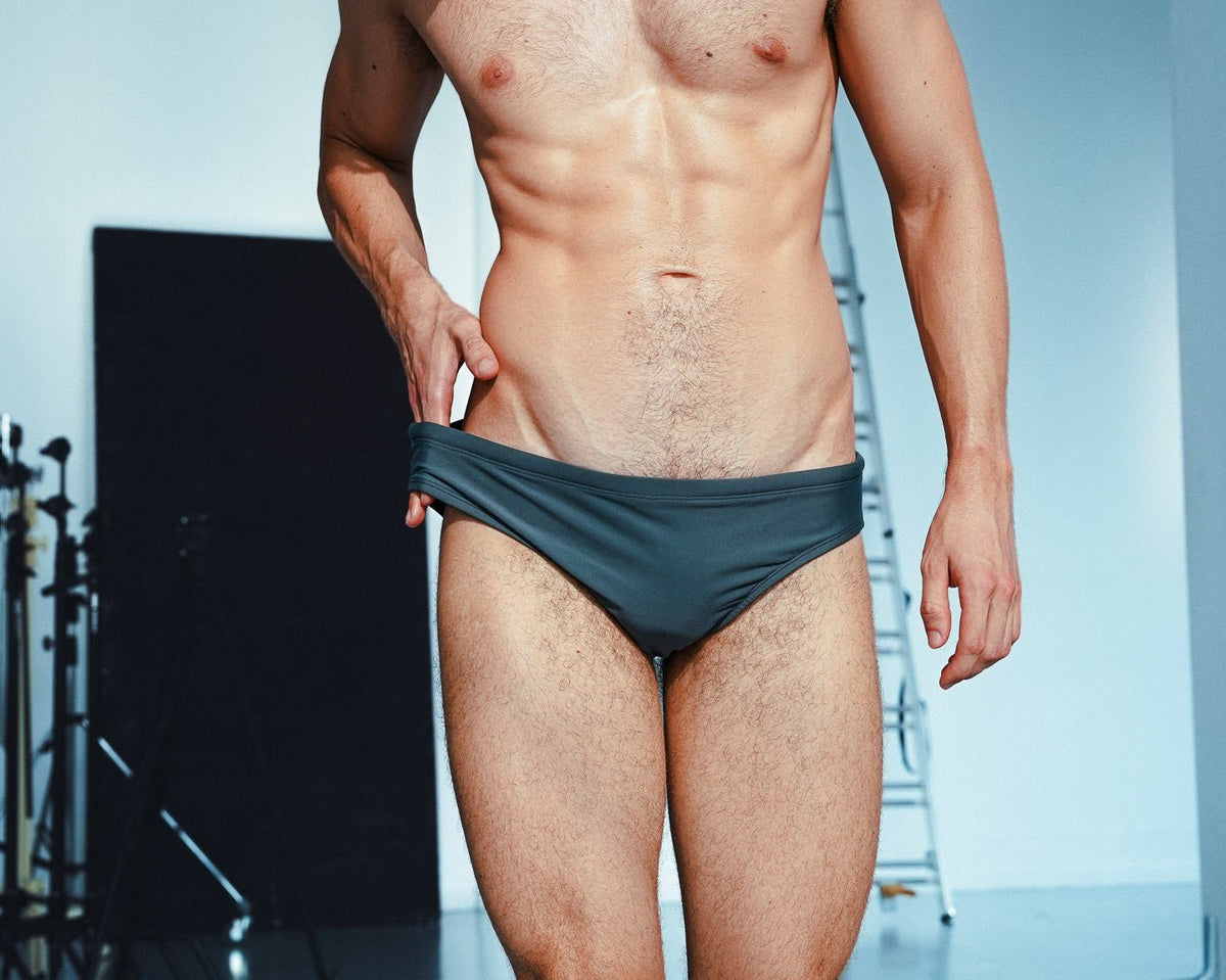 A shirtless male model wearing dark gray low-rise briefs, standing in a studio setting with a softbox and ladder visible in the background.