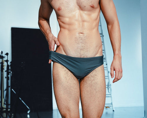 A shirtless male model wearing dark gray low-rise briefs, standing in a studio setting with a softbox and ladder visible in the background.