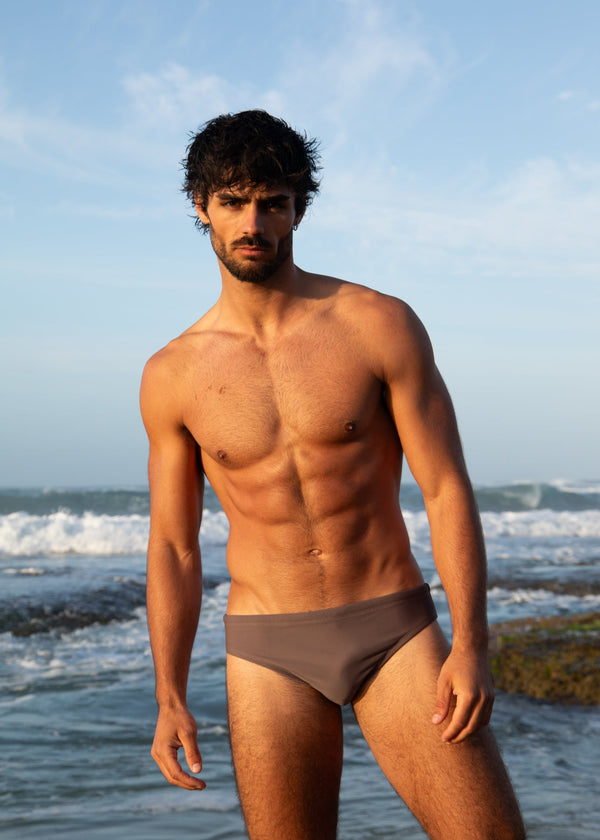 Man standing on a beach wearing gray swim trunks with ocean waves and sky in the background