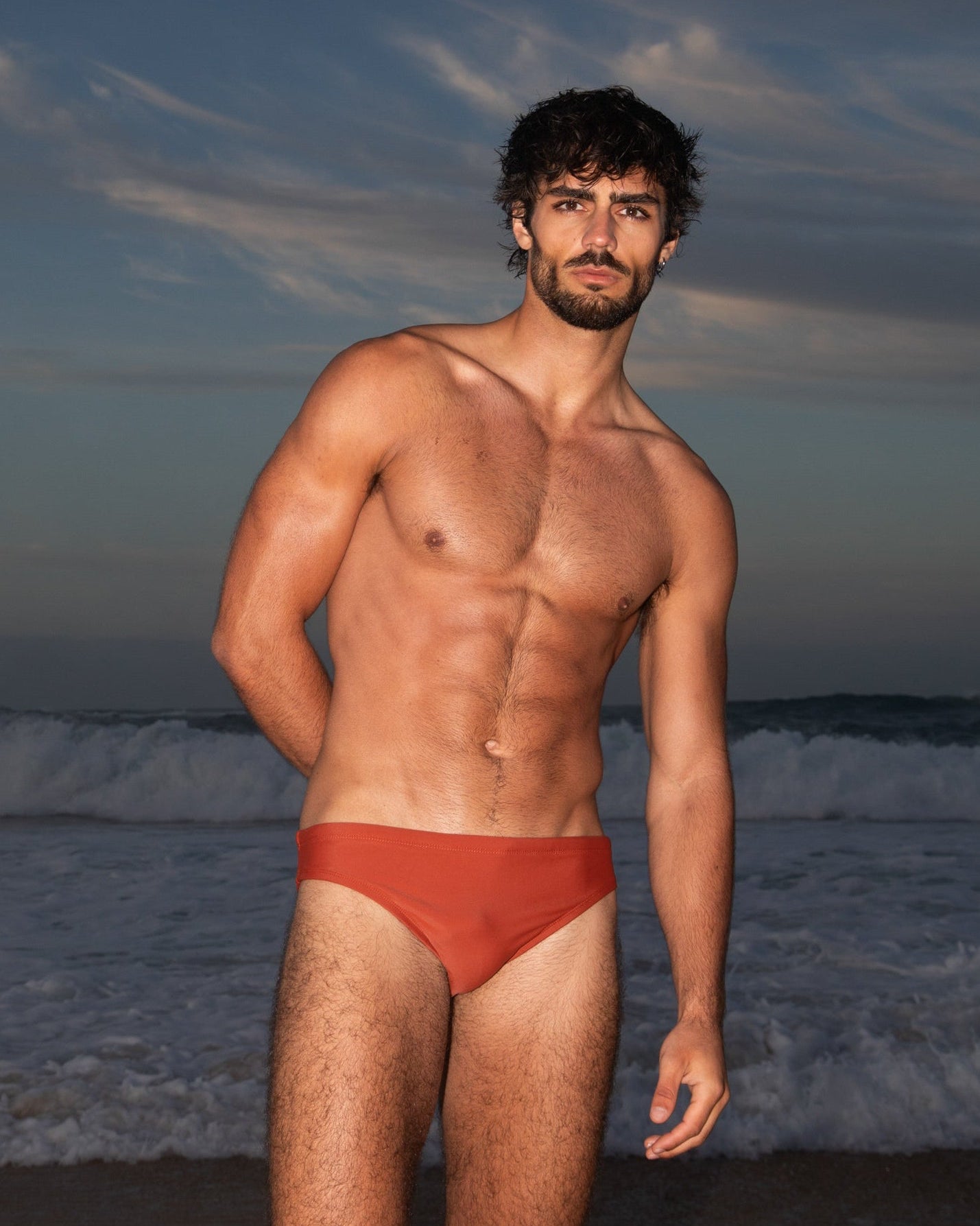 Man wearing red swim trunks standing on a beach with waves and sky in the background