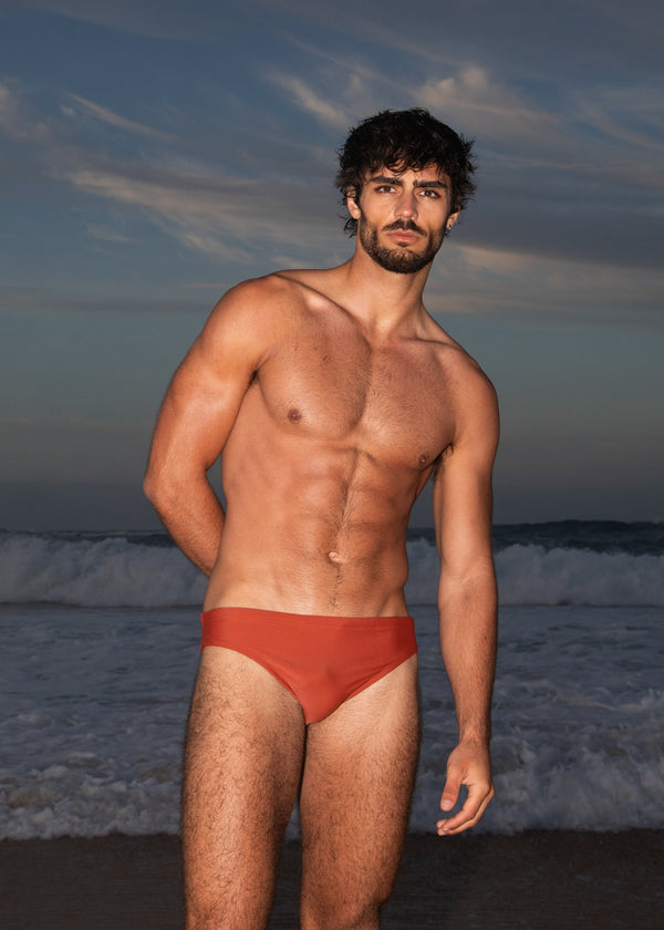 Man wearing red swim trunks standing on a beach with waves and sky in the background