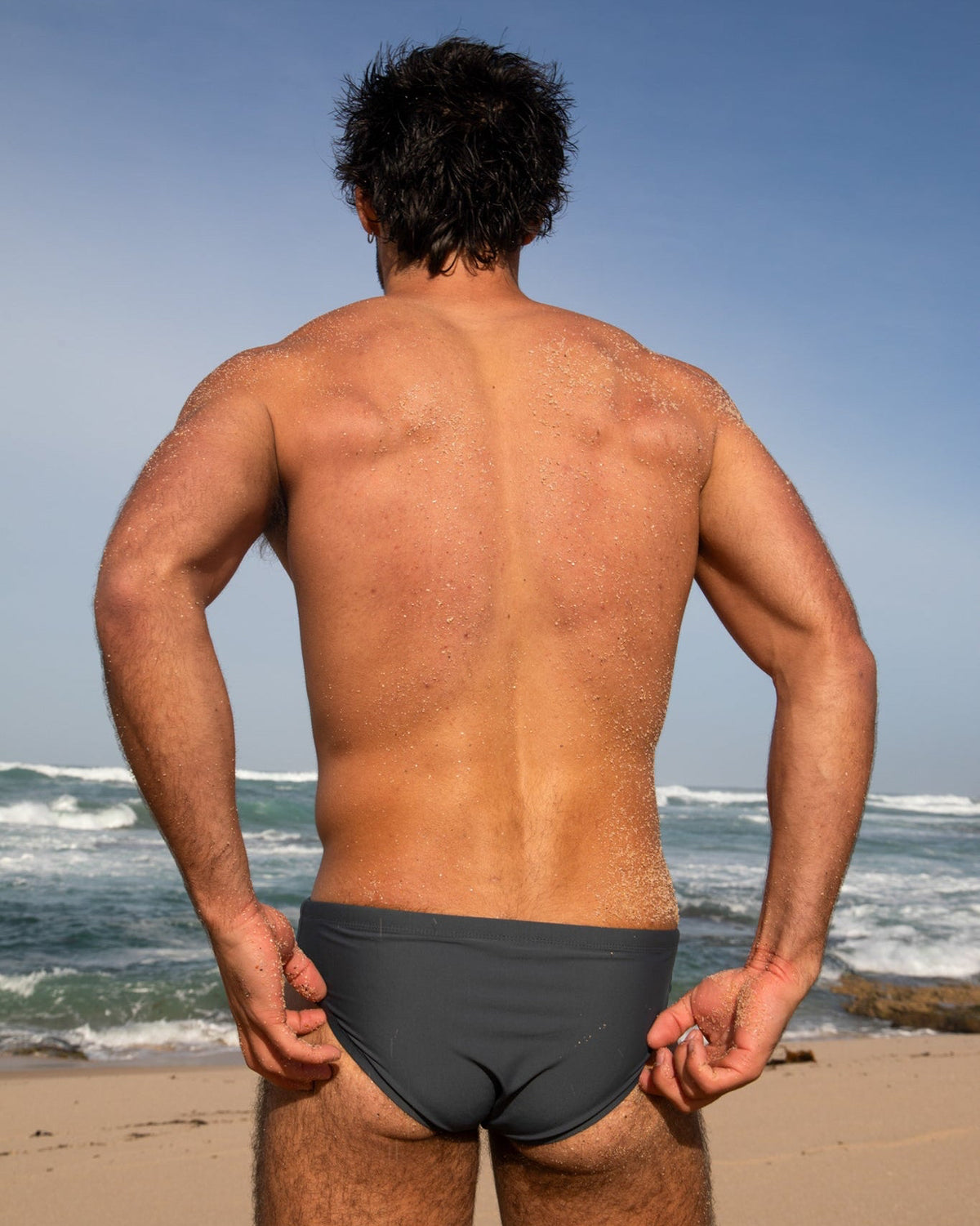 Man standing on a beach wearing black swim briefs with ocean and sky in the background