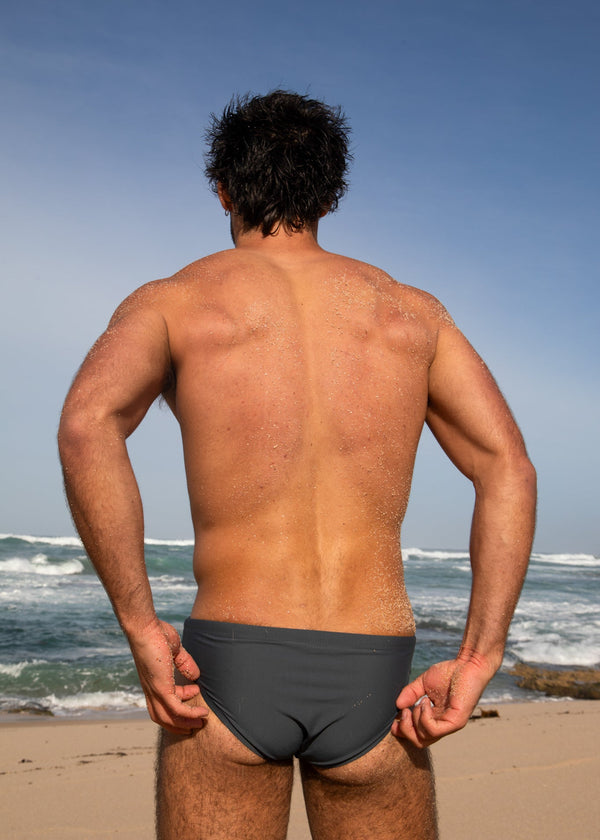 Man standing on a beach wearing black swim briefs with ocean and sky in the background