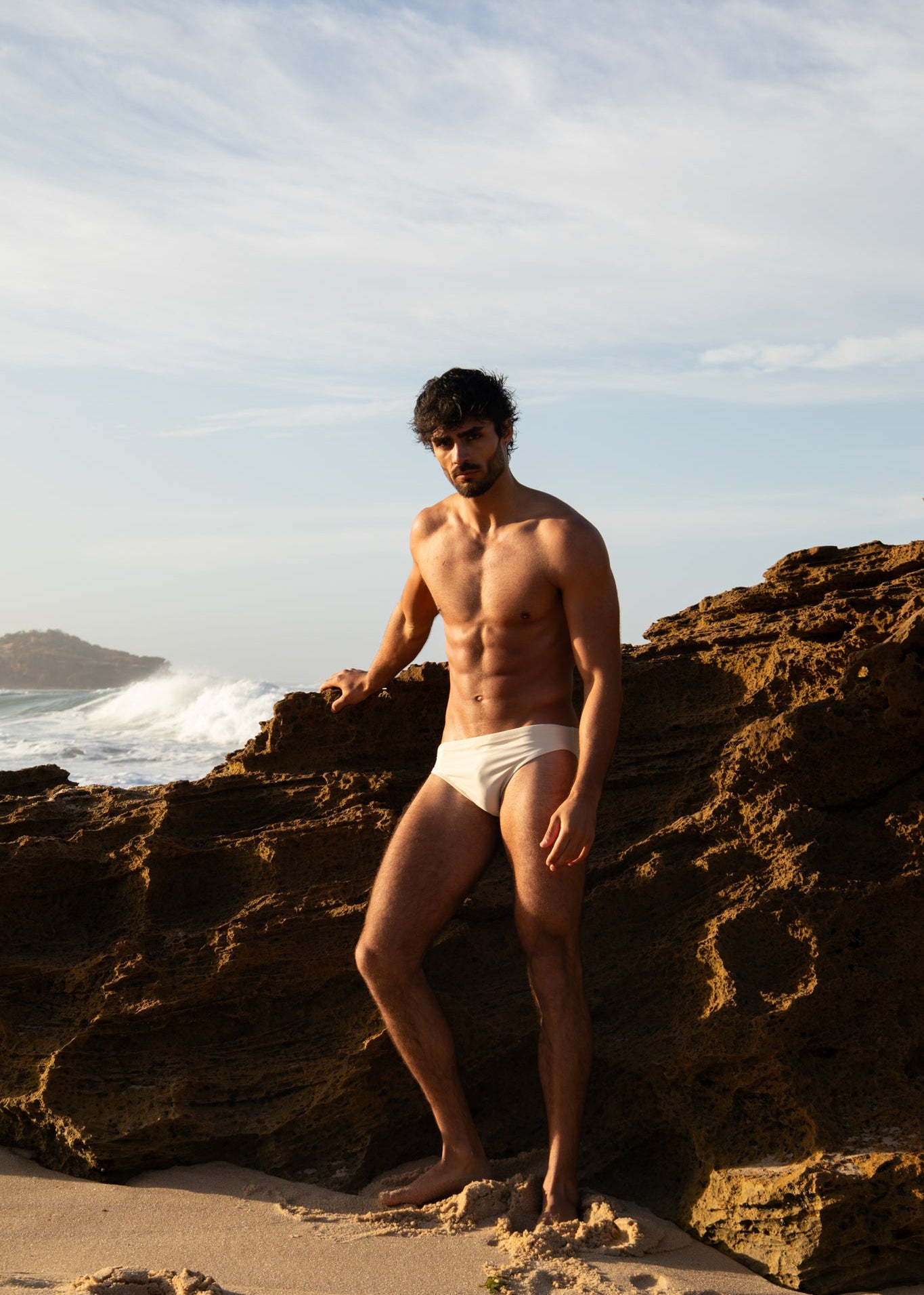 Man in white swim briefs standing on a rocky beach with ocean waves in the background