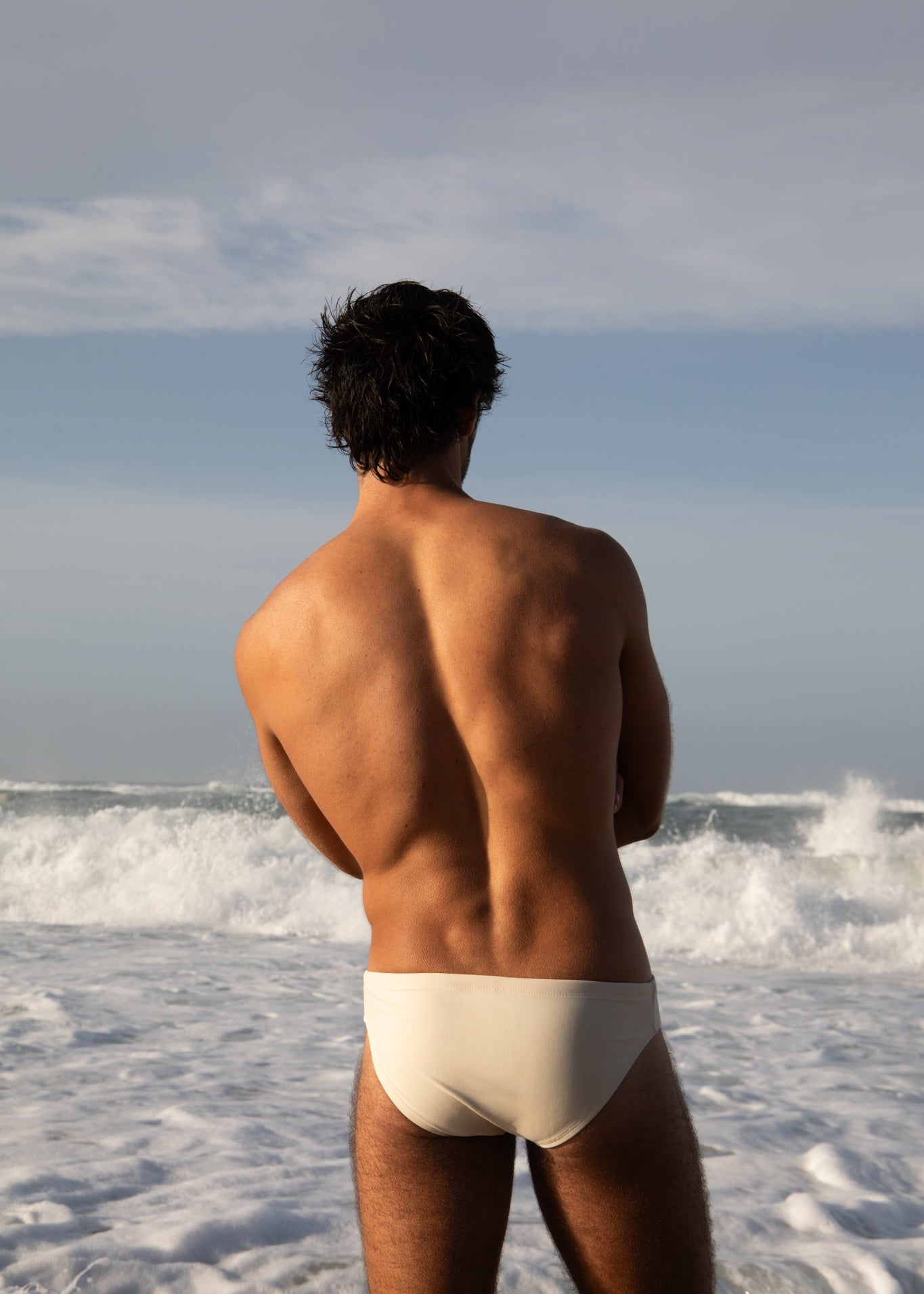 Man in white swim briefs standing on a beach with waves and sky in the background