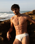 Man in white swim briefs sitting on a rocky beach with ocean waves in the background