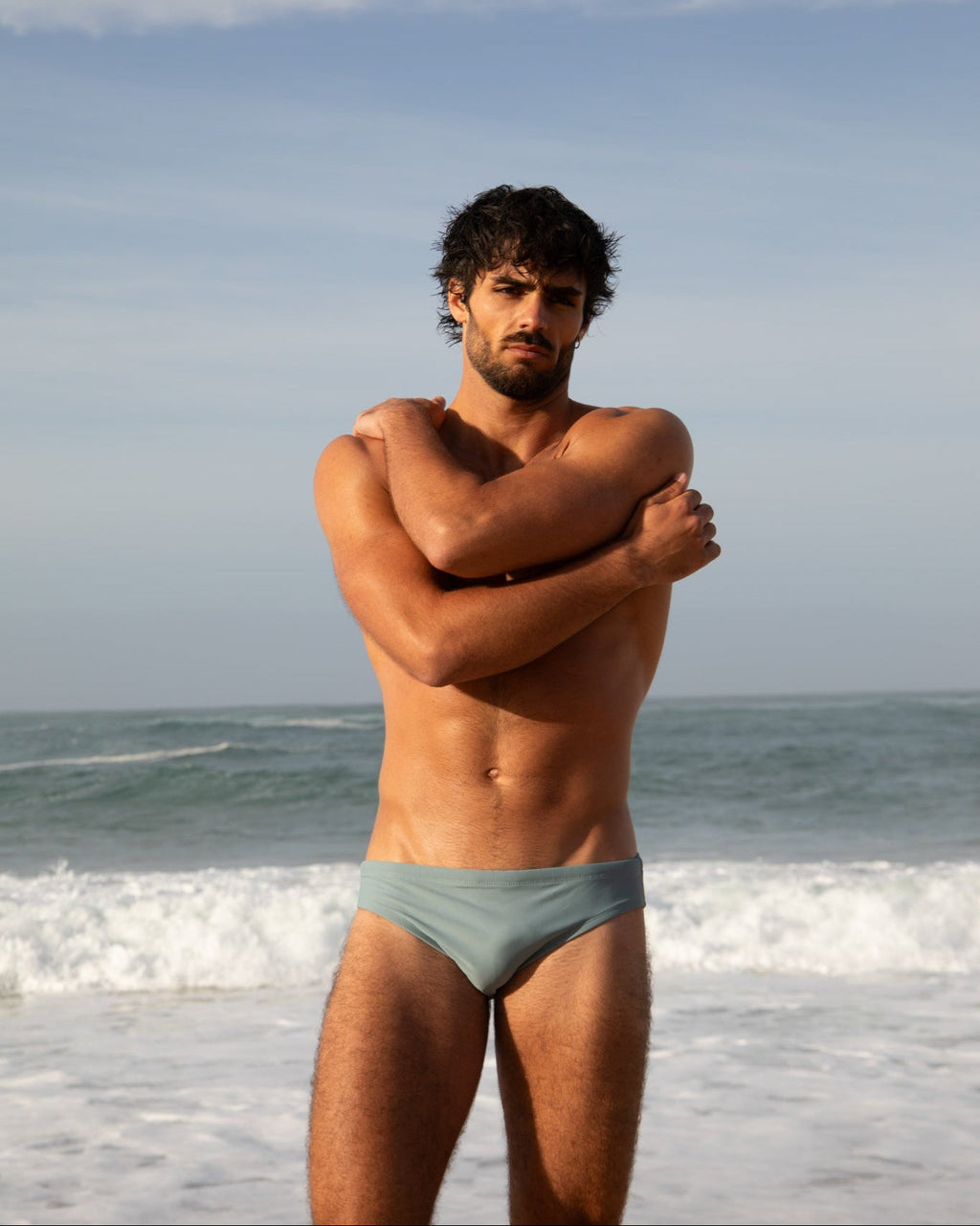 Man standing on a beach wearing light blue swim trunks with ocean and sky in the background