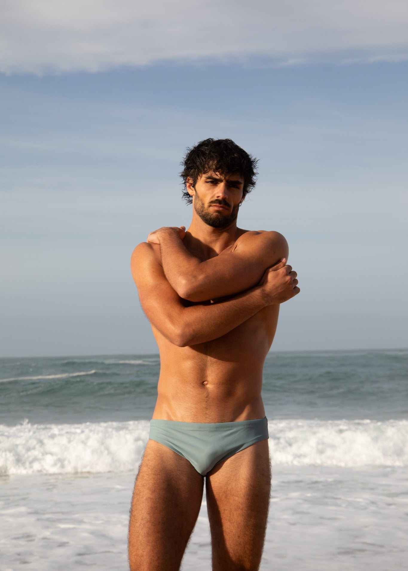 Man standing on a beach wearing light blue swim trunks with ocean and sky in the background