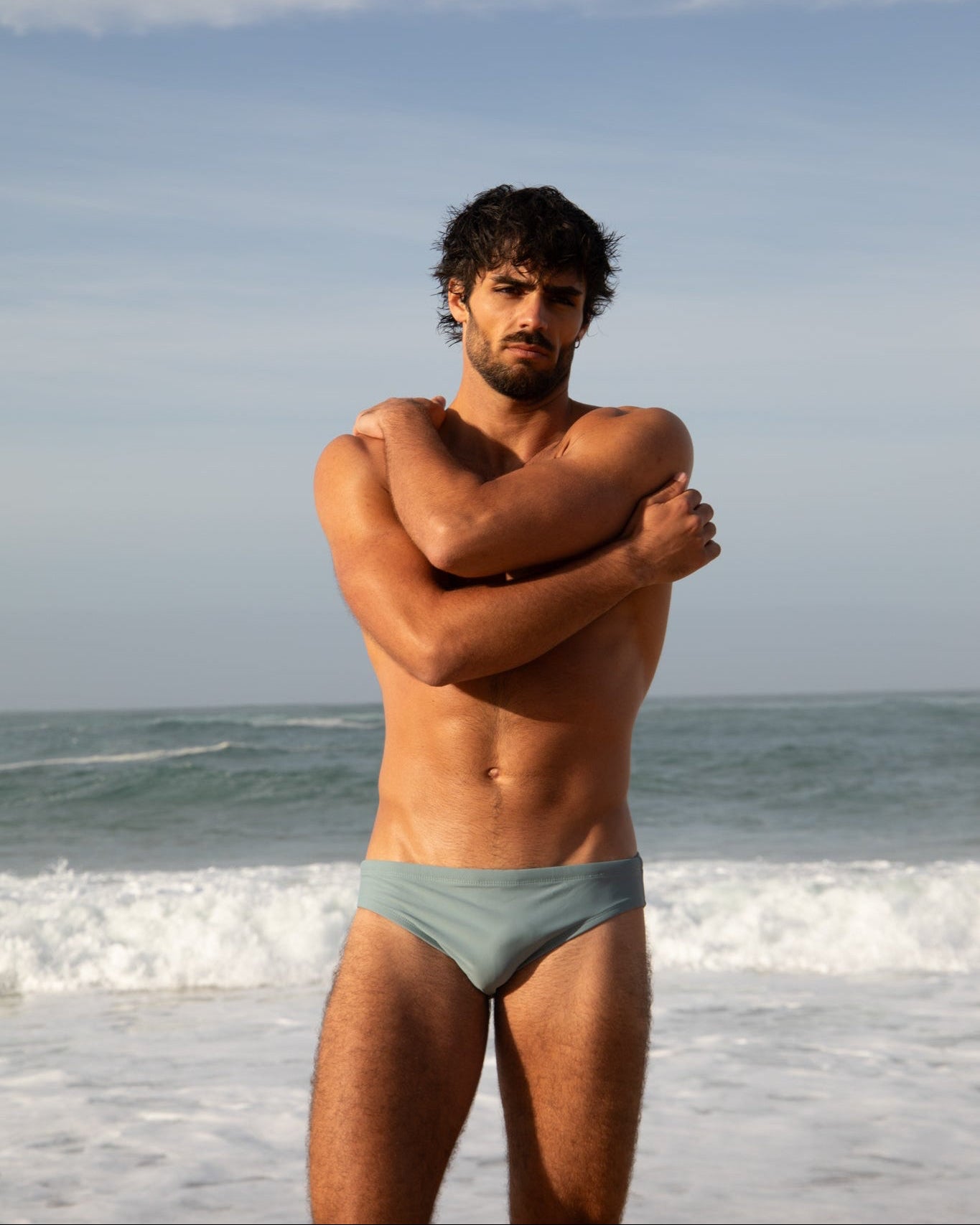 Man standing on a beach wearing light blue swim trunks with ocean and sky in the background