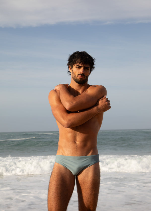Man standing on a beach wearing light blue swim trunks with ocean and sky in the background
