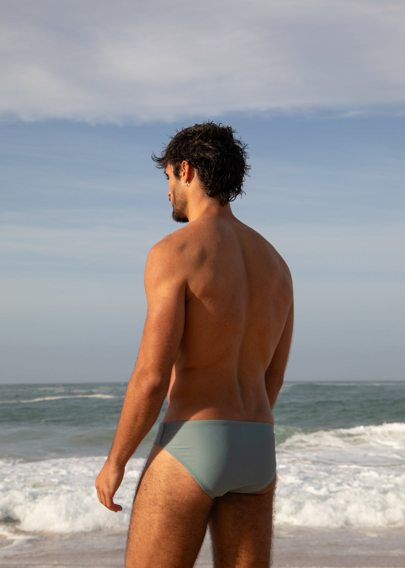 Man standing on a beach wearing a light gray swimsuit, looking out at the ocean.